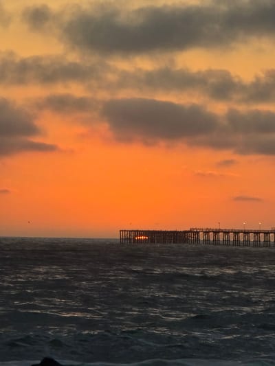 The image depicts a sunset over the ocean in Oceanside, California. A long wooden pier extends into the water from the right side of the frame, its pilings casting shadows on the dark, choppy water below. Several seagulls are perched on the railings of the pier, and a few more are in flight in the distance. The sky is a vibrant gradient of orange and yellow, with streaks of gray clouds interspersed throughout. The sun is setting behind the pier, its golden rays reflecting off the water's surface. The overall scene is one of natural beauty and tranquility.