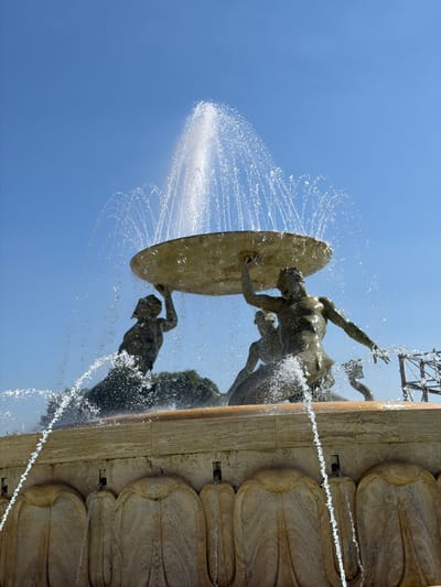 The Triton Fountain is a prominent landmark in Valletta, Malta, located just outside the city's main entrance. The fountain features four bronze tritons supporting a large bronze dish from which water cascades, creating a dynamic and impressive display. In the center of the dish, a powerful jet of water shoots upwards, creating a spectacular fountain effect against the clear blue sky. The tritons are depicted in muscular poses, straining to hold the heavy dish, adding a sense of power and movement to the sculpture. The fountain is situated on a decorative stone base adorned with classical motifs, and water streams down from the base into a pool, further enhancing the visual appeal. The overall scene captures the grandeur and artistry of this iconic Maltese monument.