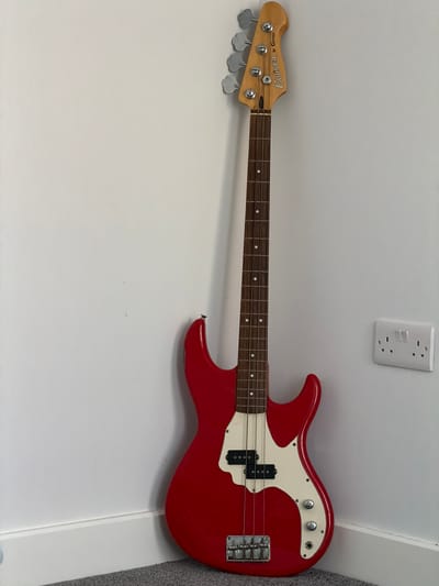 A red electric bass guitar is laying horizontally against a white wall. A white electrical outlet is visible above the guitar's body. The guitar has a white pickguard, silver knobs, and a dark-colored fretboard. The headstock has "Cruiser by Crafter" printed on it. The location in Craigavon, United Kingdom cannot be determined from the image.