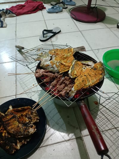 The image depicts a domestic scene in Batam, Indonesia, featuring a meal being prepared. The cooking occurs indoors on a tile floor. A metal grilling pan sits on the floor with glowing red embers inside. Raw fish coated in a yellowish-orange spice blend is arranged on a metal grill rack atop the pan, alongside skewers of meat. Another plate on the floor contains cooked, grilled fish skewers. In the background are various domestic objects: a red cloth and a variety of flip-flops, a green bowl with water, and the base of an electric fan.