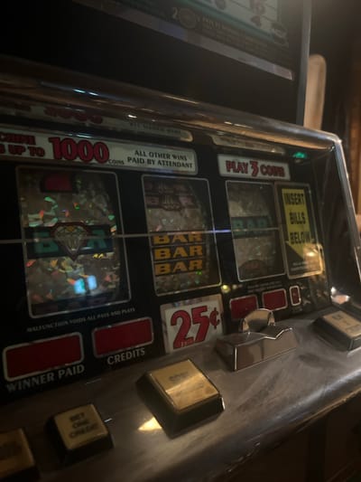 This is a close-up, eye-level shot of a slot machine located in a dimly lit, indoor environment. The machine is made of metal and plastic, with a reflective chrome surface. The focus is on the operational controls and display areas of the machine.
On the top portion of the visible machine, there is text that reads "MACHINE IS UP TO 1000 COINS". Below this, three spinning reels are visible, each displaying various symbols including a diamond and "BAR". To the right of the reels, a yellow sign reads "INSERT BILLS BELOW".
Below the reels, there are several buttons and displays. A large red button displays "25¢". To the left of this are two rectangular red displays labeled "WINNER PAID" and "CREDITS". To the right of the "25¢" display are chrome levers and buttons. One button is labeled "PLAY 3 COINS" and another is partially visible with "PLAY 1 CREDIT". There are also gold-colored buttons with text that is difficult to discern.
The overall impression is a functional slot machine, likely in operation or ready for use, in a setting that suggests a gaming establishment. The lighting is low, with some reflections on the chrome surfaces of the machine. There are no people visible in the frame, and no specific details about the surrounding environment beyond the machine itself are clearly discernible, other than what appears to be a wooden element on the right side of the frame.
The location of this slot machine in Remseck am Neckar, Germany is not determinable from the image itself.