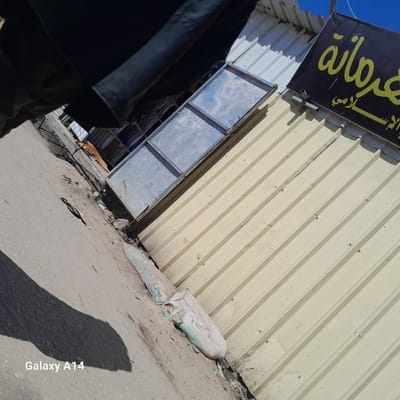 The image depicts a street scene in Khan Yunis. The foreground features a sidewalk with debris, including a torn white sack and loose trash. A partial shadow obscures the lower left corner. 

In the background, a low, one-story building stands with a corrugated metal exterior, painted a light beige. A grey metal frame structure is attached to the exterior. The building's roof is visible, constructed of corrugated metal. A sign with Arabic writing is affixed to the roof. The sky is visible in the upper corner and is a cloudless, light blue.