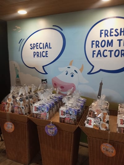 An indoor retail display is observed in Tân Kỳ Commune, Vietnam. The scene features several tall, brown, woven wicker-like baskets or bins arranged in rows, filled with merchandise. Each basket contains multiple pre-packaged product bundles, individually wrapped in clear plastic. The products include items labeled "PIE," "FRESH MILK," and other branded snacks or dairy products. Circular promotional tags are affixed to the baskets; one purple tag prominently displays "PAKET HEMAT SPECIAL 50," while an orange tag is partially visible with text including "HAPPY." The background consists of a light blue mural depicting a cartoon cow face with large eyes and pink snout, two white speech bubbles containing the phrases "SPECIAL PRICE" and "FRESH FROM THE FACTORY," and stylized silhouettes of the Statue of Liberty on the left and the Eiffel Tower on the right. Two small bird silhouettes are also part of the mural above the Statue of Liberty. Overhead recessed spotlights illuminate the display, and the ceiling appears to be a light brown, wood-toned surface. No individuals are present in the frame.