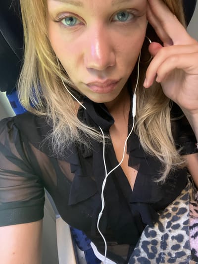 A woman with blonde hair and blue eyes is pictured. She is wearing a sheer black blouse and a leopard-print skirt. She has white earbuds in her ears. The woman is touching her head with her right hand. The background is blurry, but it appears to be an interior setting with blue seats. There is no indication of the city of Dorfen, Germany.