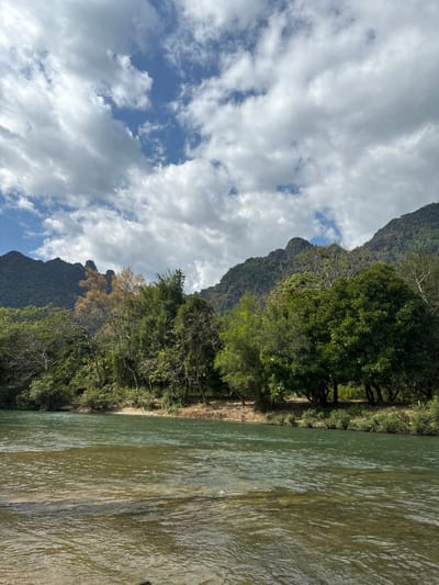 A river dominates the scene, its murky water reflecting light. The riverbank is lined with lush green trees, transitioning into rugged mountains. Fluffy white clouds fill the sky, contrasting with patches of clear blue. The landscape evokes a sense of wilderness and natural beauty.