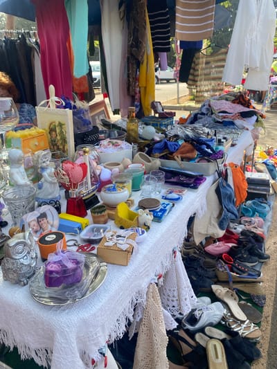 An outdoor flea market is set up on a paved area, likely a street or public square, in San Luis de Sabinillas, Spain. The foreground is dominated by a table draped with a white, fringed cloth, laden with various items for sale. These include decorative objects like a silver owl figurine, a purple heart-shaped box containing rose-shaped soaps, a framed photograph of a smiling couple, a small orange speaker, and a wooden box with ribbon. A variety of glassware, including wine glasses and tumblers, are also visible. Among the items are ceramic figurines of children, a small red boat with a checkered sail, and several glass jars. Further back on the table, a collection of small bottles, containers, and other trinkets are displayed.

Behind the table, clothing hangs from racks and a canopy structure, with dresses, shirts, and other garments in various colors and patterns. In the mid-ground, a white car is partially visible behind a pole, suggesting a roadside setting. The background reveals more stalls and a general outdoor market atmosphere, with the presence of other shoppers and vendors implied by the context of a flea market. The lighting indicates it is daytime, possibly late morning or early afternoon. The overall scene depicts a typical community market offering a diverse range of used goods and clothing.