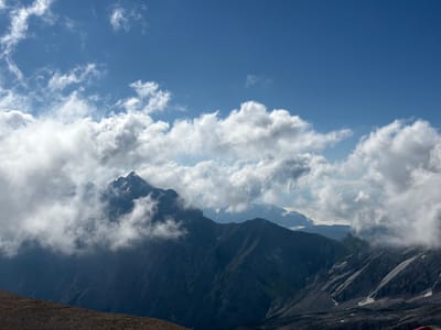 The image is a landscape view of a mountain range under a partly cloudy blue sky. The foreground shows a rocky, barren slope. In the mid-ground, dark, rugged mountain peaks rise, partially obscured by wispy white clouds. Some patches of green vegetation are visible on the slopes. The background reveals more layers of mountains, also partially covered by clouds, with a bright, clear blue sky above. The scene appears to be a natural, high-altitude environment. The image is likely taken in the vicinity of Wildermieming, Austria, given the context of the location. There are no people or man-made structures prominently visible in the image.
