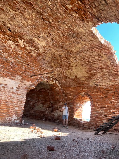 The image depicts the interior of a brick structure, likely a ruin, in Шуран, Russia. The walls are constructed of red brick and show signs of age and deterioration, with missing sections and crumbling mortar. Several arched openings are visible in the walls. A person is standing within the structure, near one of the openings. A partial view of wooden steps is present. The ground is dirt and debris. The sky is visible through openings in the structure.