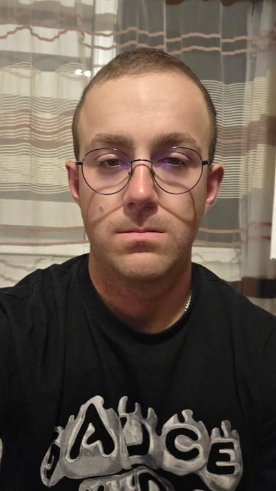 A man wearing round wire-rimmed glasses and a black t-shirt with a design is pictured from the chest up. He has short, dark brown hair and a fair complexion. The background consists of a sheer, striped curtain. The lighting casts shadows on the man's face.