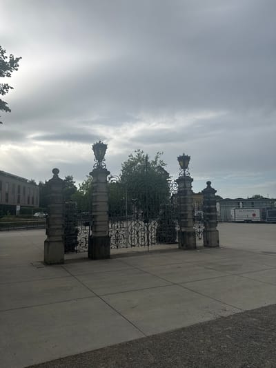 The image depicts an ornate metal gate with decorative lamps on stone pillars, likely a grand entrance. The gate is set against a backdrop of a cloudy sky and a paved plaza. To the left of the gate, a modern building with a beige facade and dark windows is visible, along with some trees. On the right side of the plaza, a white trailer with "ALTIBADI" branding is parked. The scene appears to be a public space in the city of Kreuzlingen, Switzerland, possibly near a park or a public institution, given the architectural style of the gate and the presence of a trailer suggesting an event or service. No people are visible in the frame. The overall lighting suggests an overcast day.