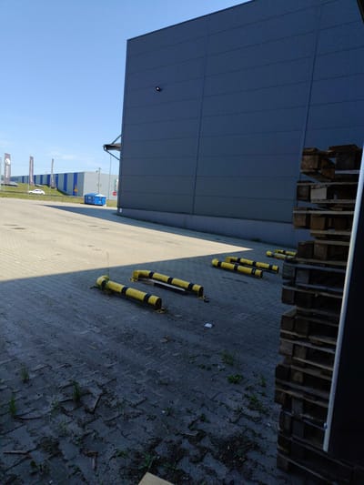 In Łomna-Las, Poland, this image captures an outdoor scene with a large, dark grey building dominating the right side of the frame. The building has a clean, modern facade composed of horizontal panels. In front of the building, a paved area made of interlocking grey bricks is visible. Several yellow and black striped bollards are positioned on this paved surface, some appearing to be protective barriers.
To the left of the building, a row of flags stands along a grassy incline. The flags display logos and text, with one clearly showing "FAGOR" and another text partially obscured by the building's shadow. Further in the background, more industrial buildings with blue and grey striped exteriors are visible under a clear blue sky. A bright blue waste bin is situated near the buildings.
A stack of wooden pallets is piled high against the side of the large grey building, casting a shadow. The paved ground is sparsely covered with patches of dirt, debris, and some weeds growing through the cracks between the bricks, indicating a somewhat neglected area. Sunlight casts sharp shadows across the pavement. No people are visible in the image.