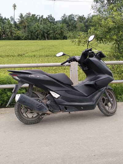 The image depicts a matte black Honda PCX 160 scooter parked on a concrete surface. Behind the scooter, a metal railing runs parallel to a vibrant green rice field. Beyond the field, a dense tropical forest with coconut trees rises in the background. The location is Uleegajah, Indonesia, indicated by the landscape. There are no people visible in the frame. The scooter is the primary subject, appearing stationary and unattended. The overall environment suggests a rural or semi-rural setting.