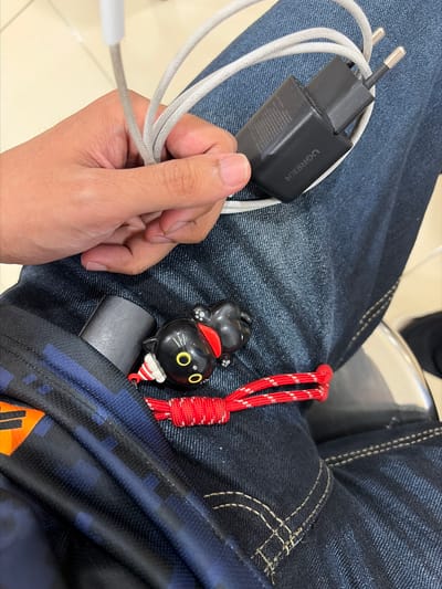 A person's hand, displaying skin with visible texture and short fingernails, holds a white charging cable connected to a black UGREEN brand charger. The charger is plugged into a black USB-A port. The cable and charger are draped over a pair of dark blue denim jeans. Partially visible in the pocket of the jeans is a cylindrical black object, possibly a vape or a battery pack. Next to this object is a small black cat-shaped keychain with yellow eyes, a red collar, and white ear details, attached to a red and white braided cord. The background is a lightly colored, textured surface, possibly a wall or floor. The scene is a close-up, focusing on the hand and the items it holds, with the jeans providing a textured backdrop. The image does not contain any elements that place it in the city of Pekalongan, Indonesia.