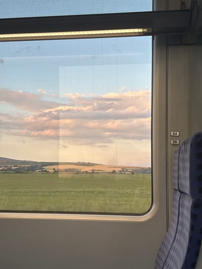 This image depicts a view from inside a train looking out a window at a rural landscape under a partially cloudy sky, with the provided context placing the scene in the city of Svoboda, Bulgaria. On the right side of the frame, a segment of a train seat is visible, upholstered in a blue fabric with a repeating pattern of light-colored, thin horizontal and vertical lines. Above the seat, two white rectangular labels with black numbering, "54" and "56", are affixed vertically to the light-colored interior wall of the train. A light fixture, featuring a grid-patterned diffuser emitting a warm yellow glow, runs horizontally above the window frame. The interior window frame is light grey with a rounded bottom edge. Faint reflections of the train's interior are visible on the window glass, including a prominent rectangular light area on the right and scattered smudges or dust particles on the upper left pane.
Through the window, the sky displays a gradient of color, from light blue at the top to soft pink and orange hues near the horizon, characteristic of twilight. Multiple altocumulus or stratocumulus clouds, primarily white with subtle pink and orange tints, are distributed across the sky. The landscape below the sky consists of agricultural fields and rolling terrain. The immediate foreground is dominated by a wide expanse of vivid green crops. Beyond this green field, the midground features a mosaic of golden-yellow fields, likely mature grain, intermingled with clusters of dark green trees and bushes. A large, verdant hill or low mountain is discernible on the far left of the horizon. The entire external scene is illuminated by soft, warm, natural light.