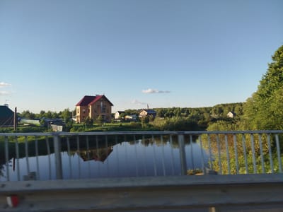 The image shows a view from a bridge in Заречье, Russia. The river reflects the houses and trees on the opposite bank. In the background, there are several houses with varied architecture surrounded by trees. A large, two-story house with a red roof is the most prominent structure. The foreground consists of a metal bridge railing. The sky is light blue with a few clouds.