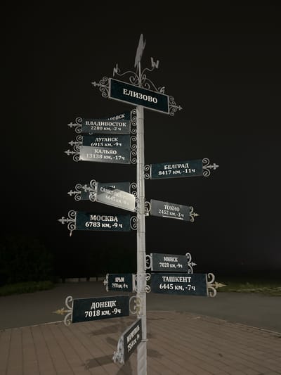 An eye-level, medium shot shows a directional signpost at night in Krasnyi, Russia. The signpost is made of metal and is painted white, with decorative swirls and filigree at the points where the signs attach. There are multiple signs pointing in various directions, indicating cities and their distances from the current location.

The signs are dark green with white lettering. From top to bottom and left to right, the visible signs read:

*   ЕЛИЗОВО (Yelizovo)
*   ВЛАДИВОСТОК (Vladivostok) 2280 км, -2 ч (2280 km, -2 h)
*   ЛУГАНСК (Luhansk) 6915 км, -9 ч (6915 km, -9 h)
*   КАЛЬЯО (Kalyo) 13138 км, -17 ч (13138 km, -17 h)
*   САНКТ-ПЕТЕРБУРГ (Saint Petersburg) 6645 км, -9 ч (6645 km, -9 h)
*   МОСКВА (Moscow) 6783 км, -9 ч (6783 km, -9 h)
*   БЕЛГРАД (Belgrade) 8417 км, -11 ч (8417 km, -11 h)
*   ТОКИО (Tokyo) 2452 км, -3 ч (2452 km, -3 h)
*   КРЫМ (Crimea) 7978 км, -9 ч (7978 km, -9 h)
*   ДОНЕЦК (Donetsk) 7018 км, -9 ч (7018 km, -9 h)
*   МИНСК (Minsk) 7020 км, -9 ч (7020 km, -9 h)
*   ТАШКЕНТ (Tashkent) 6445 км, -7 ч (6445 km, -7 h)
*   МАРШРУТ (Route) 7564 км, -9 ч (7564 km, -9 h)

At the very top of the signpost is a weathervane shaped like a bird. The background is dark, suggesting it is nighttime, with a hint of a tree on the left side and a paved area in the foreground. The paved area consists of interlocking bricks.