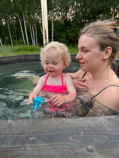 A blonde toddler in a pink swimsuit splashes water with a blue toy in a wooden hot tub. Beside the toddler, a woman with her hair in a bun looks down at the child. The hot tub is outdoors, with a forest of birch trees and greenery in the background. A silver chimney rises from behind the hot tub. The overall scene suggests a relaxed, outdoor setting in or near Ryzhkovo, Russia.