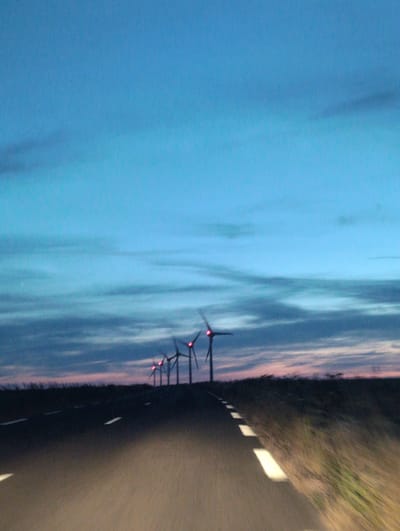 A long-exposure photograph captures a road stretching towards a line of wind turbines silhouetted against a twilight sky. The sky transitions from a deep blue at the top to lighter shades of blue and pink near the horizon, with faint clouds streaking across. The road, asphalt, has white dashed lines marking its edges and center, illuminated by the headlights of an unseen vehicle, suggesting movement. To the right of the road, tall dry grass is blurred due to motion. The wind turbines, equipped with red lights on their blades, stand in a row, their structures and blades rendered somewhat abstract by the low light and potential motion blur. The scene evokes a sense of open space and the transition from day to night.