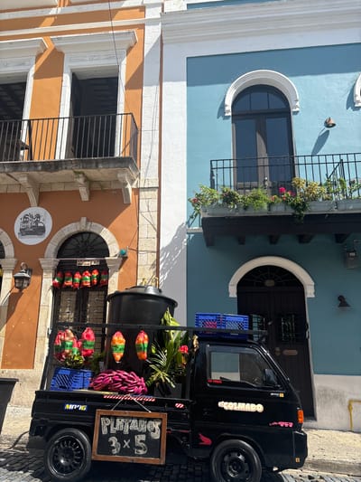 The image depicts a street scene in Old San Juan, Puerto Rico, United States of America, focusing on the colorful architecture and a parked black flatbed truck. The truck is laden with produce, including bananas and what appear to be plantains, some bagged and hanging, and others in blue crates. A chalk-written sign on the truck reads "PLATANOS 3x5". To the left, an orange building features a round sign with a black and white illustration of a truck. Red and green bags are hanging above a doorway on this building. To the right, a striking blue building rises, with an arched window and a balcony overflowing with potted plants. Both buildings exhibit architectural details common to Old San Juan, such as balconies with black metal railings and ornate window frames. The ground is paved with cobblestones, and a gas meter is visible on the exterior of the blue building. The scene is illuminated by bright sunlight, casting distinct shadows. There are no people clearly visible in the frame.