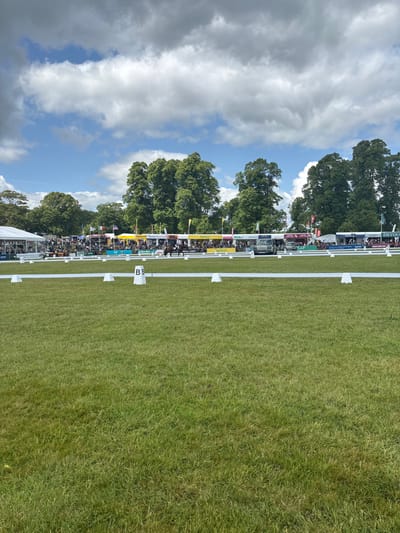The image shows a grassy field with white cones and a white tape barrier at the edge. The cones are numbered “B3”. In the distance, there are trees, tents, and a few people. The location, based on the given coordinates, is Bramham Park in West Yorkshire, United Kingdom. The weather appears to be sunny with a few clouds in the sky.  The image suggests that the area is a horse riding arena or showground.