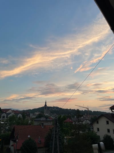 The image depicts a panoramic view of the city of Limanowa, Poland, at sunset. The sky is a vibrant canvas of blues and pinks, with streaks of orange and yellow clouds stretching across the horizon. In the foreground, the rooftops of residential buildings are visible, with a prominent church steeple rising above the cityscape in the middle ground. A construction crane is also visible, suggesting ongoing development in the area. The surrounding landscape is hilly and covered with lush greenery, indicating a natural setting within the urban environment. The overall impression is one of a peaceful, picturesque evening in a European town.