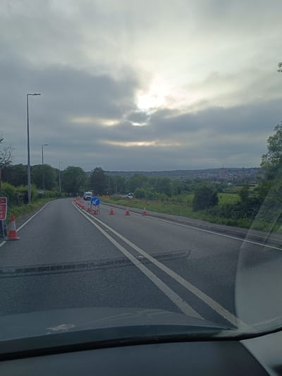 The image depicts a road with temporary traffic management in place, suggesting ongoing roadworks or an event. Orange traffic cones line the right side of the road, guiding traffic. A white van and several workers in high-visibility orange clothing are visible further down the road. A blue and white directional sign indicating a left turn is also present. The road itself is asphalt, with double white lines marking the lanes. The surrounding environment includes green verges, trees, and a distant view of a town or village with buildings nestled among rolling hills under a cloudy sky. The presence of streetlights indicates this is likely a public road. The car's dashboard and windshield are visible in the foreground, suggesting the image was taken from inside a vehicle. There is a sign on the left side of the road with red and white lettering that is partially obscured, but the word "SCHOOL" is visible. The location is identified as Arun, United Kingdom.