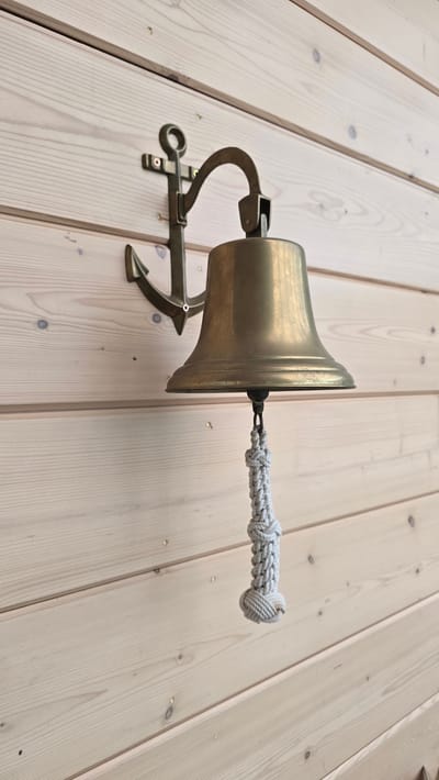 This media file features a brass bell with a rope pull, mounted on a wooden wall made of light-colored planks. The bell is designed with a classic, flared shape and a sturdy mounting bracket that incorporates an anchor motif. The rope pull is intricately braided and has a knotted end. The lighting suggests it might be daytime, with a soft, natural illumination. The wooden planks have a visible grain and occasional knots, suggesting a rustic or nautical setting. There is no visible text in the image. The scene appears to be static, with no activity taking place, focusing solely on the decorative and functional object. The overall impression is one of a well-crafted, possibly maritime-themed decorative item in a simple, natural environment.