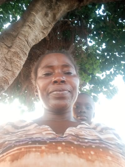 A woman poses for a selfie beneath a large tree with dense foliage. A child is visible behind her. The woman has dark hair and skin, with her shirt featuring a pattern of horizontal stripes. Sunlight illuminates the scene, creating a bright and slightly overexposed effect. The tree trunk is thick and textured, dominating the left side of the frame.