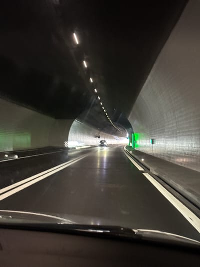 The image displays the interior of a long, concrete tunnel, captured from inside a moving vehicle with a slightly tilted perspective. The right edge of the frame reveals a dark, curved section of the vehicle's interior, likely a window frame.

The tunnel's walls and ceiling are a light gray, with striations visible, while the road surface is dark. Artificial illumination is prominent, featuring several green lights on the ceiling that project a strong glow, possibly indicating traffic signals or directional guidance. Along the left wall/roadway, a series of small, horizontally elongated white lights form a dashed line. On the right side of the dark road, two parallel white lines are painted, indicating a lane.

Further into the distance, at the end of the currently visible and well-lit section of the tunnel, two faint human figures can be seen standing or walking. A small, dark object, possibly a barrier or equipment, is also visible near them. Close to the center-right foreground, reflective white and black striped markings are on the road surface or a barrier.

The lighting within the tunnel is entirely artificial, contrasting with what appears to be daylight at the distant tunnel exit. The scene suggests travel through a modern, engineered pathway. The location context is Zurzach, Switzerland. There is no visible text in the image.