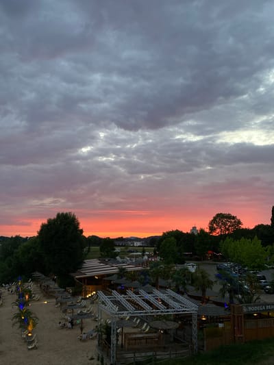 The image depicts a beach bar named "BODEGA BEACH" located in Rinteln, Germany. The scene is set at dusk, with a dramatic sunset painting the sky in vibrant shades of orange and pink. The beach area is furnished with numerous lounge chairs and umbrellas, many of which are empty, suggesting a quiet moment before the evening. Palm trees are scattered throughout the sandy area, adding to the tropical ambiance. In the background, a building with a thatched roof serves as the main structure of the beach bar, with lights visible within. Further back, trees and some distant buildings are visible, along with a parking lot occupied by several cars. The overall atmosphere is serene and picturesque.