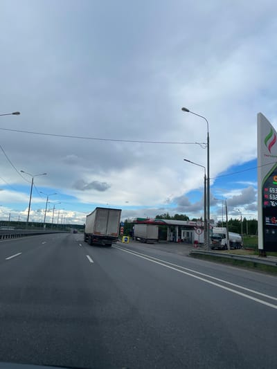 The image depicts a highway in the vicinity of a Tatneft gas station in Бавыкино, Russia. A white truck is traveling on the rightmost lane of the highway. Street lights line the road's edge. The sky is cloudy. The gas station displays fuel prices on a sign. Several other trucks are parked at the gas station.