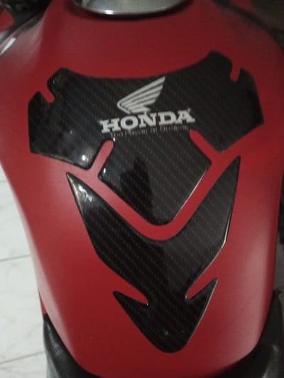 A close-up shot reveals a red motorcycle fuel tank adorned with a glossy, carbon fiber-patterned protector. This protector is shaped to fit around the Honda logo, which is prominently displayed in white. The overall design is visually striking, contrasting the red tank with the dark, textured protector. It adds a touch of sporty flair to the motorcycle's aesthetic.