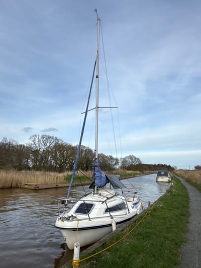 The image depicts a canal scene in North Norfolk, United Kingdom. A white sailboat, approximately 7-8 meters in length, is moored along the grassy right bank of a narrow canal. Its tall silver mast is prominent, with a furled blue mainsail cover at the boom and a blue-covered jib on the forestay. Multiple white cylindrical fenders hang along the starboard hull, resting against a wooden mooring edge. Yellow mooring lines secure the vessel to white bollards on the bank. The canal water is brown. The left bank is characterized by dense, brown reeds and a wooden platform or jetty, backed by a line of bare deciduous trees. A narrow earthen path borders the right bank. Further along the canal, a white motor cruiser with a dark canvas canopy, displaying the name "ANNA" on its stern, is visible navigating away from the viewer. The sky is overcast with scattered light clouds. No individuals are present in the visible frame. The environment is consistent with a rural waterway, typical of the Norfolk Broads within North Norfolk.