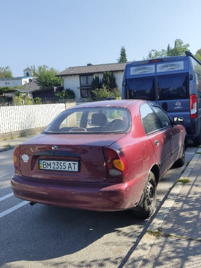 The image displays a stationary scene on a sunlit residential street in Herrsching am Ammersee, Germany. A maroon four-door Daewoo Lanos sedan, exhibiting dirt and a visibly underinflated rear left tire, is parked on the asphalt pavement, oriented away from the viewer. The vehicle bears a Ukrainian license plate, "BM 2355 AT," and a white circular sticker on its trunk lid. Directly behind the sedan, a blue Fiat Ducato van, featuring "Eco Royal" branding on its rear windows, is also parked. To the left of the vehicles, a white wooden picket fence separates the street from a property, beyond which several residential buildings with brown tiled roofs and green trees are visible. In the distant background, a white church tower with a green dome, consistent with the St. Nikolaus Church, rises above other structures. A concrete sidewalk with intermittent grass growth borders the street on the right. Strong shadows from the vehicles and overhead trees indicate a clear, sunny day. No individuals are present within the frame.