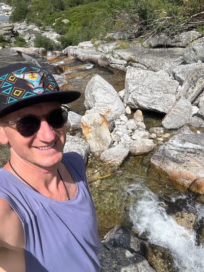 This is a portrait-style, high-angle, close-up selfie of a person standing near a rocky stream. The person is wearing a black baseball cap with colorful geometric patterns and an embroidered design of an animal's head, and round-lensed sunglasses. They are also wearing a light purple t-shirt and a black cord necklace. The person is smiling broadly at the camera. The background consists of large, grey rocks and a fast-flowing, clear stream with whitewater. Green vegetation is visible on the left side of the image, suggesting a natural, outdoor setting. The lighting indicates it is daytime, likely sunny given the bright conditions and shadows. The rocks and water suggest a mountainous or alpine environment. There is no visible text in the image. The overall mood is cheerful and adventurous.