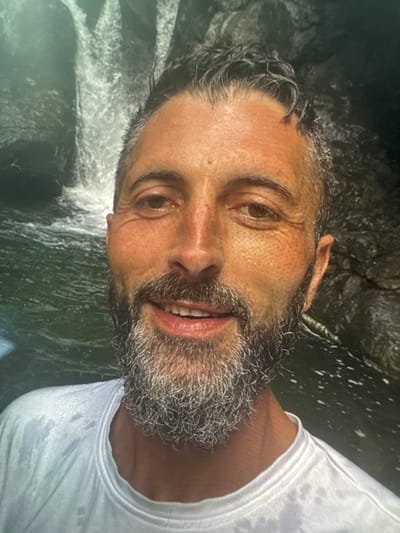 A male individual is captured in a close-up portrait, positioned in front of a natural waterfall and a body of water, located in the city of Gennevilliers, France. The man, appearing middle-aged, has short, wet, dark hair with visible graying and a full, wet, gray beard. He looks directly at the camera with a slight smile, his eyes dark brown. He wears a wet, white, short-sleeved t-shirt. The background consists of a cascading waterfall over dark rock formations, with a pool of water visible at the base and to the sides. The overall environment suggests a natural, outdoor setting.