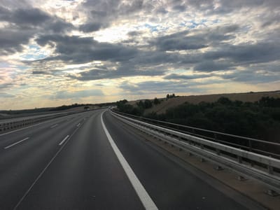The image depicts a highway in Klipphausen, Germany, on a cloudy day. The perspective is from a moving vehicle, showing the asphalt road with white lane markings curving to the right. A metal guardrail runs along the right side of the road, separating it from a grassy embankment and trees. In the distance, there are fields and scattered trees, with wind turbines visible on the horizon. The sky is filled with dramatic, grey clouds, with patches of sunlight breaking through. There are no people or other vehicles clearly visible in the immediate foreground.