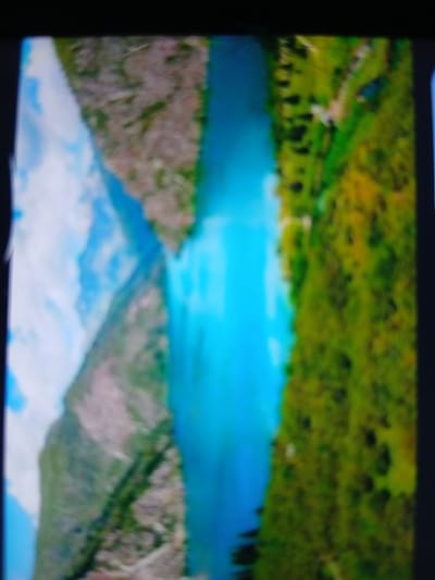 This image is a distorted aerial view of a landscape. A bright turquoise river or body of water runs through the center, bordered on the left by a steep, rocky mountainside with some vegetation, and on the right by a lush, green, forested hillside. The sky is visible on the far left, appearing cloudy and bright blue. The overall impression is of a natural, mountainous region, possibly a canyon or valley. The specific location in the city of Auk, Kyrgyzstan, cannot be determined from this image alone, as no urban features or landmarks are visible.