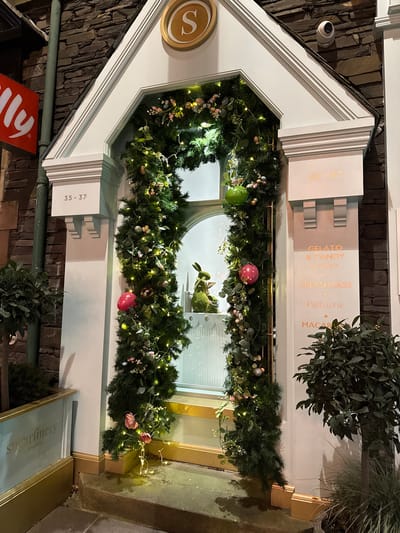 The image depicts the window display of the Sugarinery in Windermere, United Kingdom, as it is decorated for Easter. The building's exterior is made of dark stone. A white frame surrounds the window, with signage indicating the address as "35-37." The window display features a green moss bunny surrounded by greenery, Easter eggs, and fairy lights. Above the display, text advertises "Gelato & Candy Parlour," "Milkshakes," "Patisserie," and "Macarons." A security camera is visible on the building's exterior. An Illy sign is partially visible beneath the window.