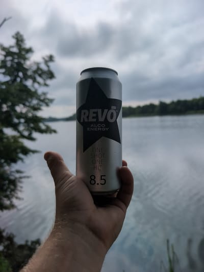 A person's hand holds a silver Revo alcohol energy drink can in front of a body of water and a cloudy sky. The can has black text and a black star graphic. The text on the can reads "REVO", "ALCO ENERGY", "ONE SHOT ONE HIT", and "8.5". The background shows a lake or river with trees on the far bank under a gray, overcast sky. This scene is not located in the city of Perliavka, Ukraine.