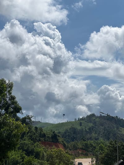The image captures a hilly landscape with lush green vegetation under a partly cloudy sky. In the foreground, trees frame the scene on the left and right. A paved road is visible in the middle ground, with a few pedestrians and a vehicle. Power lines stretch across the undulating hills in the background. The sky is dominated by large, billowy white and gray clouds, interspersed with patches of blue. The overall impression is a serene, natural environment with signs of human infrastructure. It's not possible to determine the specific location within Xayden, Laos, from the provided image.