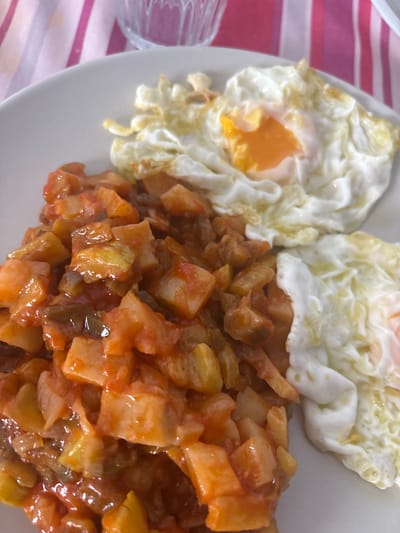 The image presents a directly overhead, slightly angled view of a meal served on a white, round plate. The plate rests on a vertically striped fabric tablecloth, featuring alternating wide magenta and narrow white bands. On the left side of the plate, a portion of diced, cooked vegetables, predominantly yellowish-brown cubes resembling potatoes, is coated in a glossy, reddish-orange sauce, with darker vegetable pieces interspersed. To the right, two fried eggs are visible. The upper egg displays a bright orange, semi-liquid yolk encircled by a crispy, white, and ruffled albumen. The lower egg shows a similar white and ruffled albumen. In the upper left background, a portion of a clear, textured glass is visible out of focus. The setting is an indoor dining environment located in Urbanización Encinares De Alcolea, Spain.