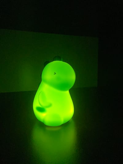 A bright green, dinosaur-shaped night light glows in a dark room. The light is positioned on a dark, reflective surface, casting a green glow upwards onto a textured, dark green wall. The night light has a rounded body, a small head with a subtle eye marking, and two small arm-like protrusions on its sides. The lighting is dim, with the night light being the primary source of illumination in the scene. The overall impression is one of a child's room or a decorative item placed in a dimly lit area. The image does not contain any information that would place it in Limanowa, Poland.