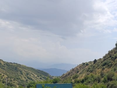 This is a wide-angle, eye-level, outdoor, daytime photograph taken in Saylyk, Uzbekistan. The image depicts a valley landscape with rolling hills covered in green vegetation and scattered trees. The hillsides are dry and appear to be arid, with patches of brown grass and rocky terrain. In the background, several layers of mountains are visible, fading into a hazy, muted blue. The sky is overcast with soft, diffused clouds. In the foreground, the edge of a dark teal, corrugated metal roof is visible, suggesting a building or structure is present at the bottom of the frame. There are no people or animals visible in the image. The overall scene is one of natural, undeveloped mountainous terrain.