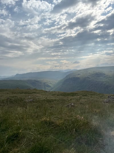 A scenic landscape photograph depicts a mountainous vista. The left side of the image showcases a sky filled with elongated, grey clouds. To the right, a grassy slope descends into a valley, revealing distant, tiered mountain ranges veiled in a faint haze. The color palette is dominated by muted greens, greys, and blues, conveying a serene and somewhat overcast atmosphere. The angle of the photo is slightly tilted.