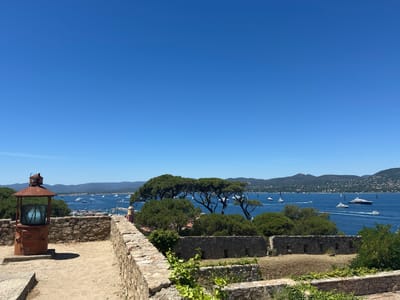 This is a wide-angle, eye-level, outdoor, daytime photograph capturing a scenic view of the bay of Saint-Tropez, France, from a raised vantage point, likely within the citadel. The foreground is dominated by a rustic, sandy path and a weathered stone wall. To the left, a vintage, rusty red lighthouse-like structure with a glass lens stands prominently. The middle ground features lush green trees, including a cluster of pine trees with their characteristic umbrella-like canopies, and more stone structures that appear to be part of ancient fortifications. Beyond the trees, the deep blue water of the bay is dotted with numerous boats, including large yachts and sailboats, all moving. In the distance, a lush, green, and hilly coastline with scattered buildings stretches across the horizon under a clear, bright blue sky. There are no people visible in the image. The overall impression is one of a tranquil, sunny day overlooking a busy maritime scene.