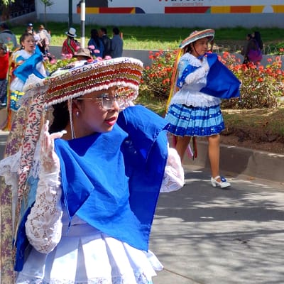 In the city of Wanchaq, Peru, two young girls participate in a parade. The girl in the foreground, on the left, wears a traditional outfit with a white dress and a highly decorative hat adorned with beads and colorful trim. She holds a bright blue fabric in her hands and wears white lace gloves. To her right, another girl, also in traditional attire, wears a white and blue dress with a striped skirt and a hat. She holds a blue cloth. Both girls are walking on a paved surface, with a background of green grass, bushes with red flowers, and other parade participants. The scene is lit by sunlight, casting shadows on the ground.