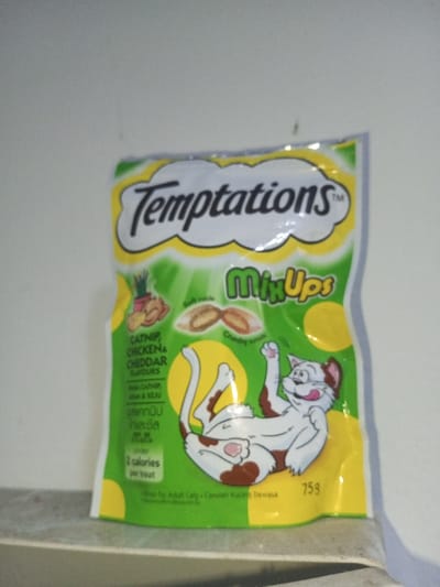 The image shows a bag of Temptations MixUps cat treats with catnip, chicken, and cheddar flavors. It features a cartoon cat lying on its back. The bag also indicates the treats have soft and crunchy centers and contain 2 calories per treat. The packaging includes text in another language. The net weight of the treats is 75g.