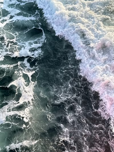 The scene, located in the coastal waters off Kato Karioutes, Greece, displays an overhead view of agitated ocean water. The image is bisected diagonally by a large, turbulent wave crest, predominantly white with areas exhibiting faint pinkish-purple coloration in its foam, dominating the right side. To the left, the water is a dark teal-green, textured by numerous smaller, irregular patterns of white seafoam and surface ripples. No individuals, vessels, or man-made objects are present within the frame. The environment is exclusively marine, characterized by natural wave action and water movement.