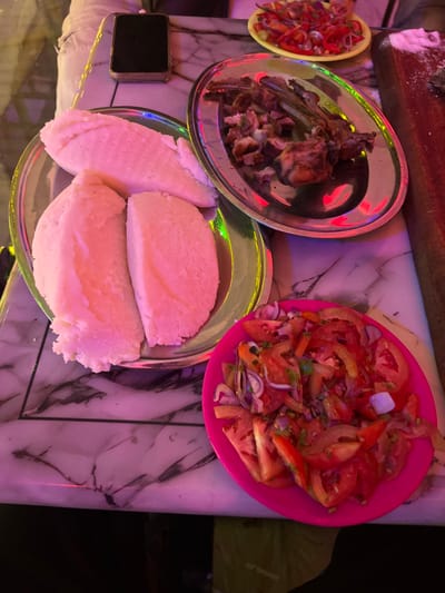 The image presents a top-down view of a meal laid out on a table, likely in Kimorori, Kenya, during the evening or night, indicated by the artificial pink and green light reflections on the surfaces. The table itself has a glossy, marble-patterned white surface.
The main subjects are various dishes common in Kenyan cuisine. A large silver-colored metal plate holds generous portions of a white, starchy food, which appears to be ugali. Next to it, another silver-colored metal plate contains pieces of grilled or roasted meat, possibly goat or beef ribs and chunks.
Two salads, locally known as kachumbari, are also part of the spread. One is served in a bright pink plastic plate/bowl and consists of sliced red tomatoes and red onions, mixed with what appears to be green herbs. A smaller, yellow plastic plate/bowl with a similar tomato and onion salad is partially visible in the upper left corner.
Other objects on the table include a dark, rectangular item, possibly a mobile phone or wallet, situated to the left of the ugali plate. A wooden surface with some white powder, possibly salt or spices, is visible in the top left background.
No people are visible in the image, and there is no discernible text. The scene depicts a meal prepared for consumption, characterized by its local culinary elements and the atmospheric artificial lighting.